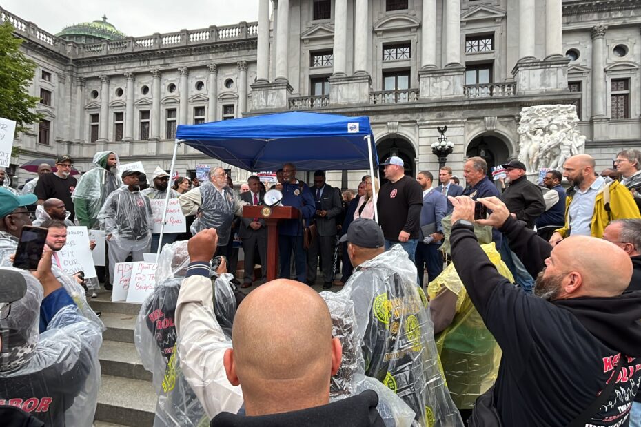 Color image of a crowd of public transportation workers surrounding a tent with a podium with a Black man in glasses speaking, on the steps in front of the Pennsylvania state capitol building in the rain.