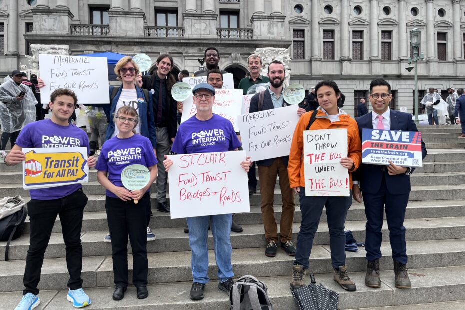 Color image of a multiracial, multigender group fo people on the steps of the Pennsylvania state capitol holding signs supporting public transportation. Signs on the edges read "Transit For All PA!" and "Save the Train"