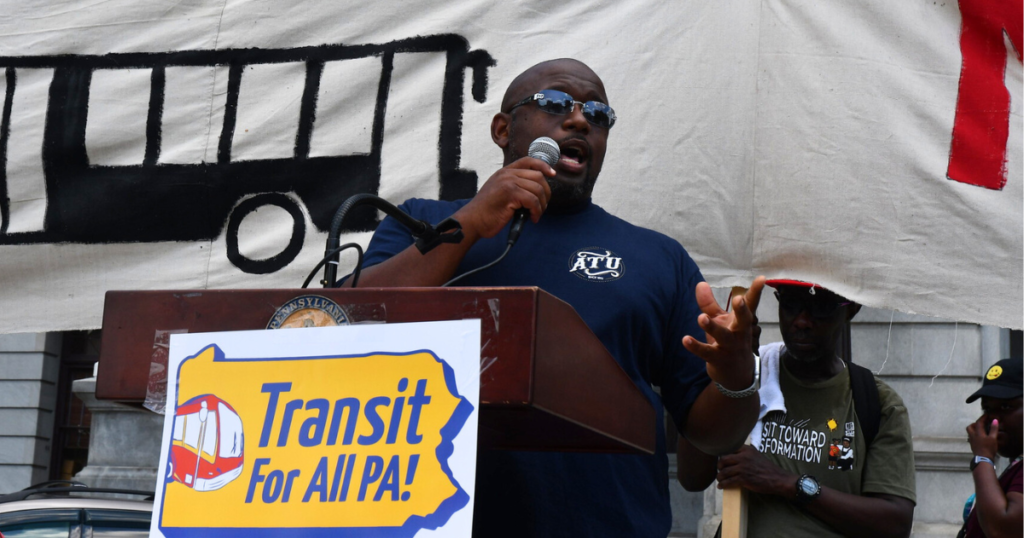 image description: photo of Lionel Randolph speaking at Transit for All PA! rally in Harrisburg. He wears a blue Amalgamated Transit Union shirt at a podium that has a Transit for All PA! sign. He's standing in fron of a large hand-painted banner with a bus on it.