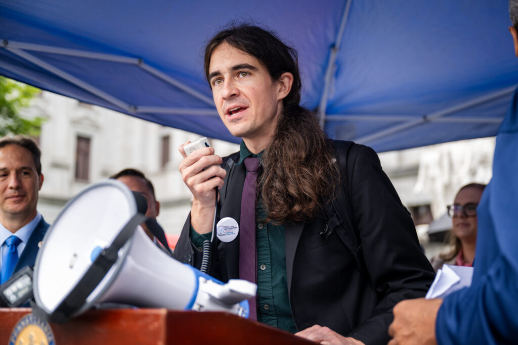 Color image of a white person with a ponytail under a tent, speaking at a podium through a megaphone wearing a green shirt under a black jacket, with a maroon tie