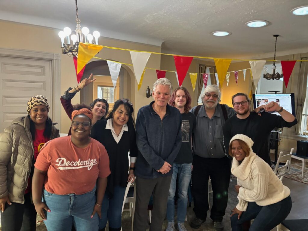 Color image of a group of people smiling of mixed races, ages, and genders, under yellow, red and white flags in a room together