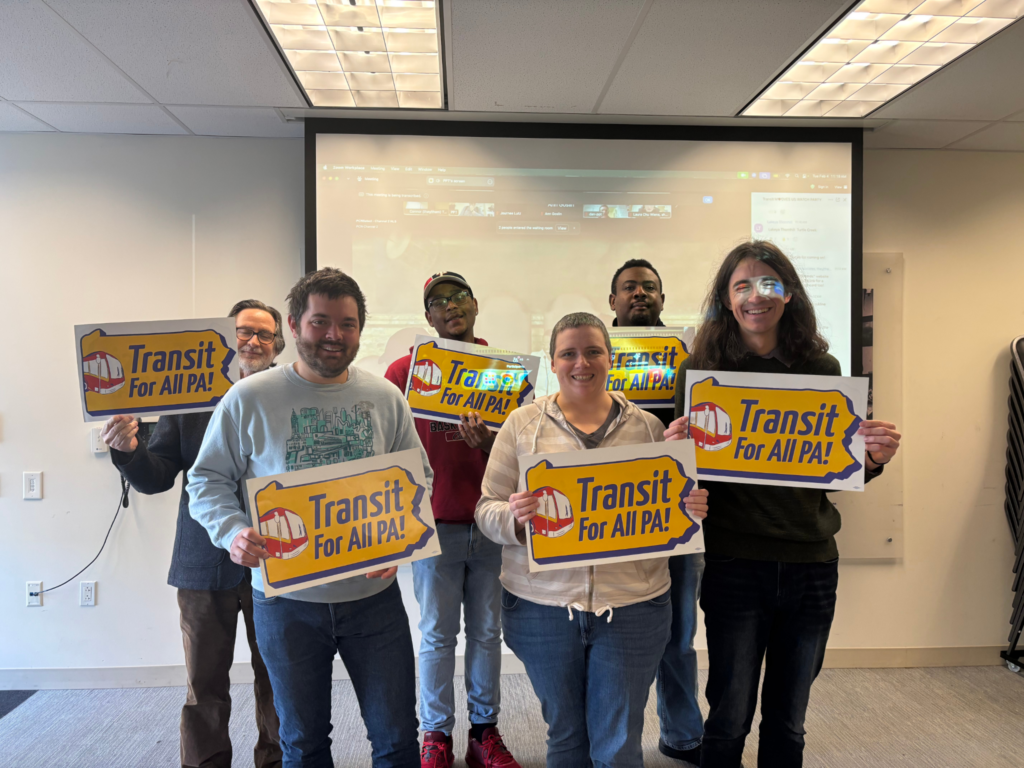 Color image of a smiling group of six people of different genders, races, and ages smiling and holding yellow and red signs with white backgrounds in the shape of Pennsylvania reading "Transit For All PA!" with a red cartoon bus